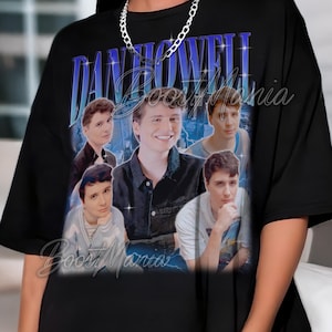 Puede incluir: Camiseta negra con un diseño gráfico de estilo vintage que presenta a una persona con el nombre "DAN HOWELL" en azul y morado. La camiseta incluye varias imágenes de la persona. El texto "Boost Mania" también es visible.