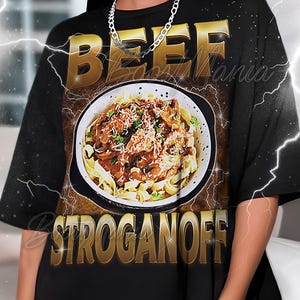 Könnte beinhalten: Schwarzes T-Shirt mit einem Aufdruck von Beef Stroganoff in einer weißen Schüssel. Die Wörter "BEEF" und "STROGANOFF" sind goldfarben, schwarz umrandet. Das T-Shirt hat eine lockere Passform.
