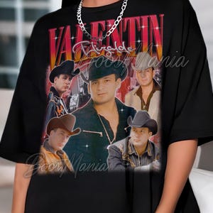 May include: Black t-shirt featuring a vintage-style graphic of Valentin Elizalde with the text "Valentin Elizalde" in red and gold. The shirt includes multiple images of the singer and the text "Boos Mania".