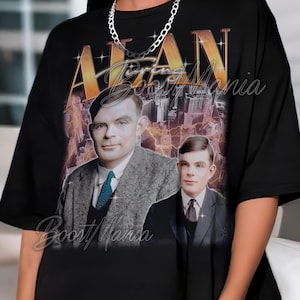 May include: Black t-shirt featuring a portrait of Alan Turing with the text "Alan Turing Boost Mania" in gold. The design includes two images of Turing and a cityscape background. A silver chain necklace is worn.