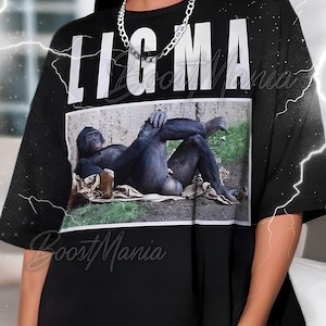 Puede incluir: Camiseta negra de gran tamaño con la palabra "LIGMA" en letras blancas grandes. La camiseta presenta una imagen rectangular de un chimpancé tumbado en el suelo. Se lleva un collar de cadena plateada alrededor del cuello.