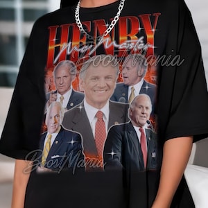 May include: Black t-shirt featuring a graphic design with the name "HENRY" in red and orange, with multiple images of a man in suits. The text "Boost Mania" is also visible.