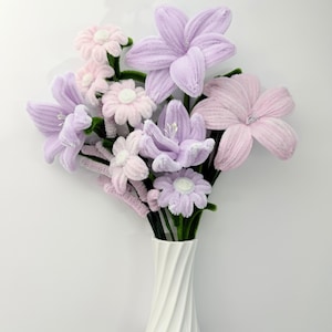 May include: A bouquet of handmade flowers in a white vase. The flowers are in shades of lavender and pink, with green stems. The vase has a twisted design.