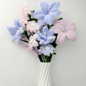 Può includere: Un bouquet di fiori fatti a mano in un vaso bianco. I fiori sono un mix di azzurro e rosa, con steli verdi. Il vaso ha un design a spirale.