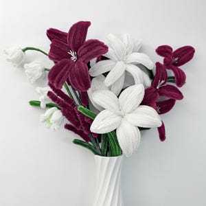 Può includere: Un bouquet di fiori fatti a mano in un vaso bianco. L'arrangiamento presenta fiori bordeaux e bianchi con steli verdi. Il vaso ha un design a spirale. I fiori sembrano essere fatti di un materiale morbido e vellutato.