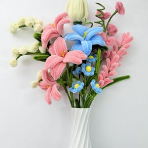 Può includere: Un vivace bouquet di fiori fatti a mano in un vaso bianco. La composizione presenta fiori rosa, blu e gialli con steli verdi. Il vaso ha un design unico e attorcigliato, che aggiunge un tocco moderno all'esposizione floreale.