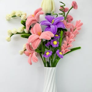 Peut inclure: Un bouquet de fleurs fait à la main dans un vase blanc. Les fleurs sont faites d'un matériau doux et duveteux dans les tons de rose, de violet, de jaune et de blanc. Le vase a un design torsadé.