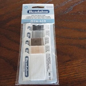 May include: A package of Beadalon 100% silk bead cord in various neutral colors. The package includes five 2-meter (6.5 ft) spools of cord, ideal for jewelry making. The label includes the text "100% Silk Bead Cord".