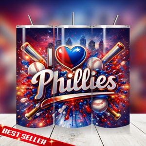 May include: A stainless steel tumbler featuring a vibrant design with the text "I ❤️ Phillies." The design includes baseballs, bats, and a heart-shaped emblem in red, white, and blue, set against a cityscape background. The words "Best Seller" are also visible.