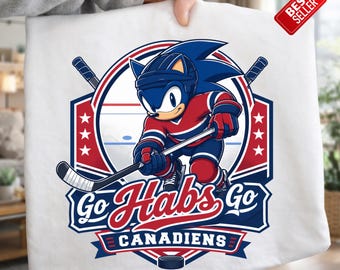 Puede incluir: Camiseta blanca con un dibujo de Sonic the Hedgehog con uniforme de hockey, sosteniendo un palo de hockey. El diseño incluye el texto "Go Habs Go Canadiens" en rojo y azul, con un disco de hockey en la parte inferior. Una etiqueta de "Más vendido" está adjunta.