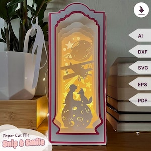 May include: A paper cut file for a 3D layered lamp with a pink and white frame. The design features a silhouette of a boy and a fox on a moon with a plane flying overhead. The text "Snip & Smile" is at the bottom of the image.