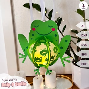 Könnte beinhalten: Eine grüne Papierschnittdatei eines Frosches, der eine leuchtende Laterne mit einem Froschdesign hält. Die Datei ist für ein Papierbastelprojekt und in den Formaten AI, DXF, SVG, EPS und PDF verfügbar. Der Text "Snip & Smile" befindet sich am unteren Rand des Bildes.