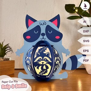 Könnte beinhalten: Blaue Papier-Raccoon-Lampe mit einem weißen und schwarzen Waschbärgesicht auf der Vorderseite. Die Lampe ist kugelförmig und hat ein ausgeschnittenes Design aus Blättern und einem Waschbärgesicht. Die Lampe steht auf einer Holzoberfläche.