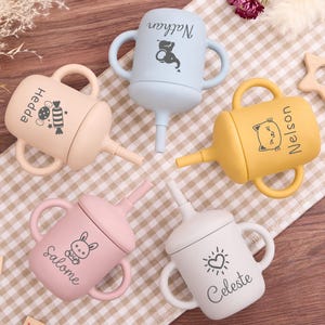 May include: Four colorful baby sippy cups with handles and spouts. Each cup is a different pastel color: beige, blue, yellow, pink, and white. Each cup has a name and a small graphic printed on it.