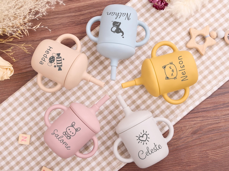May include: Four colorful baby sippy cups with handles and spouts. Each cup is a different pastel color: beige, blue, yellow, pink, and white. Each cup has a name and a small graphic printed on it.