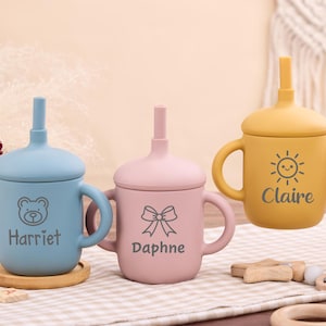 May include: Three silicone baby sippy cups in blue, pink, and yellow. Each cup has a lid with a straw and handles. The blue cup has a bear graphic and the name "Harriet". The pink cup has a bow graphic and the name "Daphne". The yellow cup has a sun graphic and the name "Claire".
