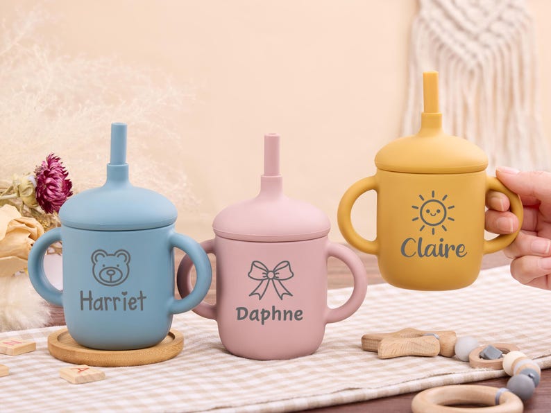 May include: Three silicone baby sippy cups in blue, pink, and yellow. Each cup has a lid with a straw and handles. The blue cup has a bear graphic and the name "Harriet". The pink cup has a bow graphic and the name "Daphne". The yellow cup has a sun graphic and the name "Claire".