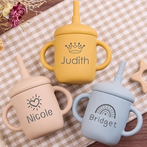 May include: Three silicone baby sippy cups in yellow, peach, and blue. Each cup has a handle, a lid with a straw, and a name printed on the front: Judith, Nicole, and Bridget. A wooden star-shaped teether is also visible.