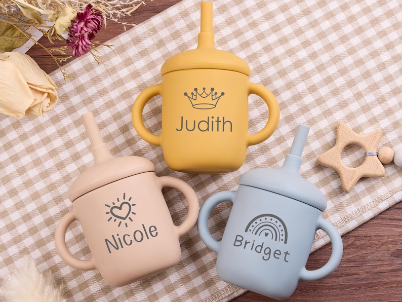 May include: Three silicone baby sippy cups in yellow, peach, and blue. Each cup has a handle, a lid with a straw, and a name printed on the front: Judith, Nicole, and Bridget. A wooden star-shaped teether is also visible.
