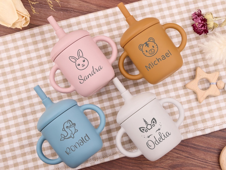May include: Four colorful silicone sippy cups with handles and straws. Each cup is a different color: pink, tan, blue, and white. The cups are decorated with animal illustrations and names: a bunny, a tiger, a dolphin, and a unicorn.