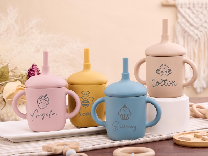 May include: Four colorful baby sippy cups with lids and handles. The cups are pink, yellow, blue, and tan. Each cup has a different design and name: a strawberry and "Angela", a bee and "Nicol", a cupcake and "Sidney", and a monkey and "Colton".