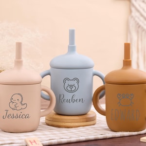 May include: Three personalized baby sippy cups in beige, blue, and brown. Each cup features a different animal graphic and a name: Jessica, Reuben, and Edward. The cups have handles and a straw.