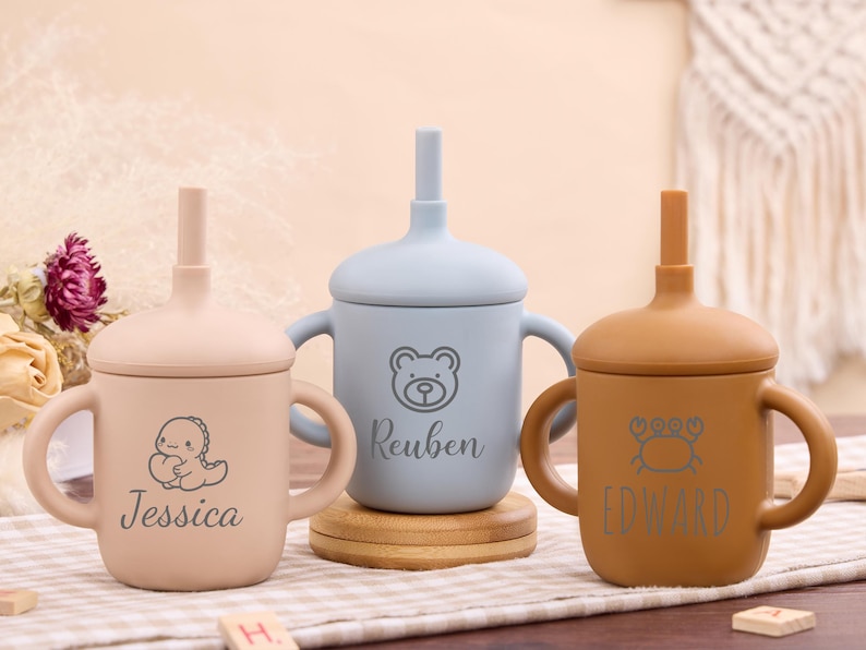 May include: Three personalized baby sippy cups in beige, blue, and brown. Each cup features a different animal graphic and a name: Jessica, Reuben, and Edward. The cups have handles and a straw.