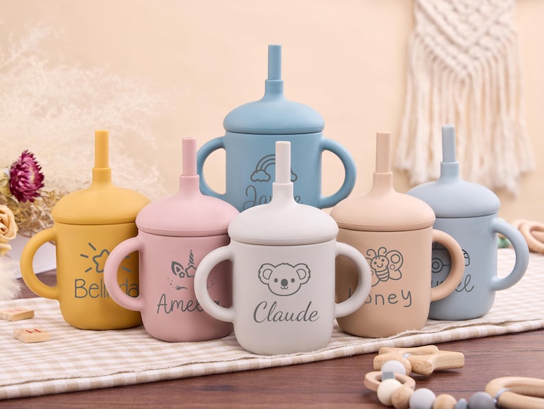 May include: A collection of colorful silicone sippy cups with handles and attached straws. Each cup is a different pastel color, including yellow, pink, blue, and beige. The cups are decorated with various designs and names, such as a sun, a unicorn, a koala, and a butterfly.