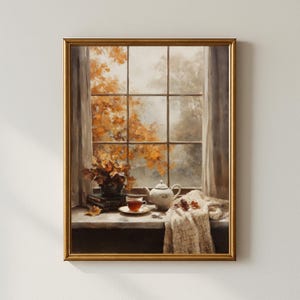 Puede incluir: Una pintura enmarcada representa una escena otoñal. Una ventana con vistas al follaje otoñal es el telón de fondo de una naturaleza muerta. Una tetera, una taza de té y una manta descansan en el alféizar. Un jarrón de flores y libros completan la cálida composición.