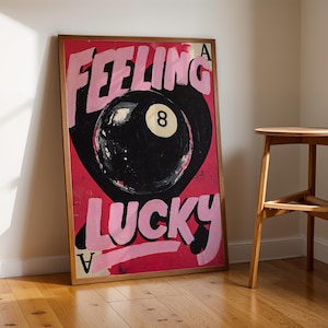 May include: A framed art print with the words "Feeling Lucky" in pink over a black 8-ball on a red background. A wooden stool is to the right of the artwork, and the artwork is leaning against a white wall.