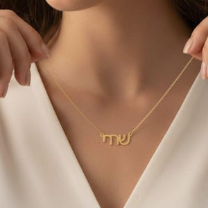 Puede incluir: Un collar dorado con un colgante que deletrea una palabra en escritura hebrea. El collar se lleva alrededor del cuello. El fondo es una blusa blanca.