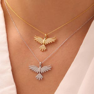 May include: Two necklaces with dove pendants. One necklace is gold-toned, the other is silver-toned. The dove pendants have outstretched wings and are adorned with small, sparkling embellishments. The necklaces are displayed against a white shirt.