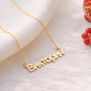 May include: A gold-colored necklace with the name "Виктория" in Cyrillic script. The necklace is on a white cloth, with a red Matryoshka doll and red berries in the background. The necklace is a piece of jewelry.
