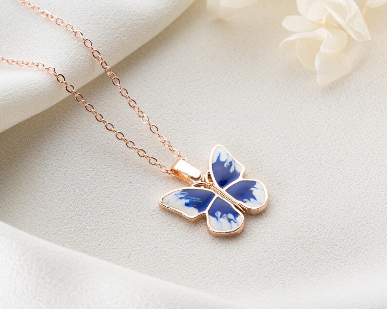 May include: A delicate rose gold chain necklace featuring a blue and white butterfly pendant. The butterfly has a marbled design and is a statement piece.