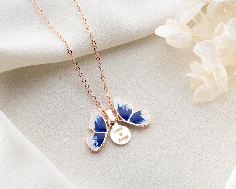 May include: A rose gold necklace featuring a blue and white butterfly pendant. The pendant has a small round charm inscribed with the words "Love Always".