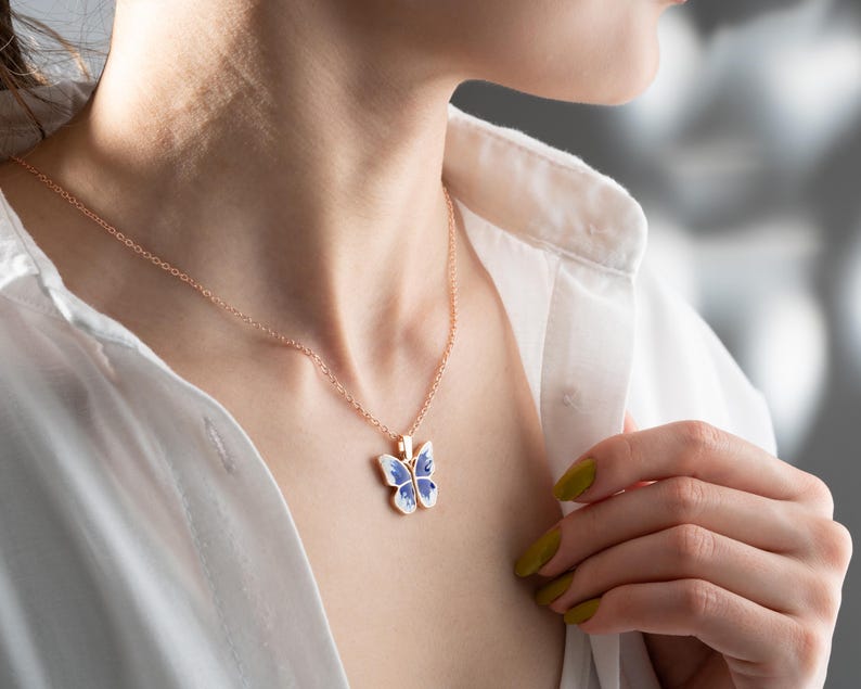 May include: A delicate gold chain necklace with a blue and white butterfly pendant. The butterfly has a detailed design and is a beautiful piece of jewellery.
