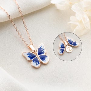 May include: A rose gold necklace with a blue and white butterfly pendant. The pendant has a small round charm with the text "Love is Always" engraved on it.