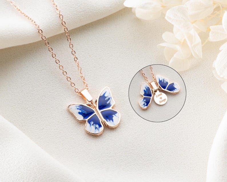 May include: A rose gold necklace featuring a blue and white butterfly pendant. The pendant has a small round charm with the text "Love is Always" engraved on it.