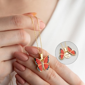 Könnte beinhalten: Goldfarbene Halskette mit Schmetterlingsanhänger mit roten und schwarzen Emaille-Details. Der Schmetterlingsanhänger hängt an einer zarten Goldkette. Die Halskette ist ein Schmuckstück.