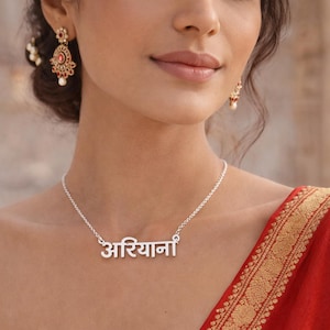 May include: A silver necklace with the name "अरियाना" in Hindi script. The necklace is worn with matching earrings and a red sari with gold trim. The image is a close-up of the jewelry.