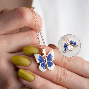 May include: A delicate gold chain necklace featuring a blue and white butterfly pendant. The butterfly has a white body and blue wings with a white swirl pattern. The pendant is attached to a small round charm engraved with the words "Love is Always".