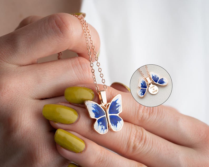 May include: A delicate gold chain necklace featuring a blue and white butterfly pendant. The butterfly has a white body and blue wings with a white swirl pattern. The pendant is attached to a small round charm engraved with the words "Love is Always".