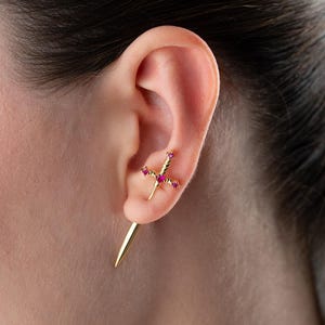 Può includere: Un orecchino dorato a forma di spada con una lama appuntita rivolta verso il basso e un'impugnatura decorata con tre piccole pietre preziose rosa brillante. L'orecchino è inserito nel lobo, con l'impugnatura che poggia contro la cartilagine.