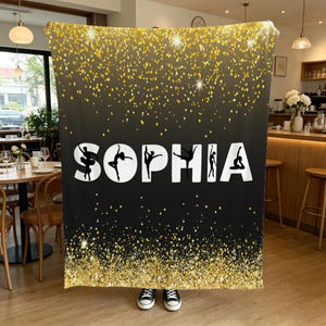 May include: A rectangular throw blanket featuring a black background with a gold glitter gradient effect at the top and bottom edges. The name SOPHIA is printed in large white capital letters, with black silhouettes of dancers inside each letter.