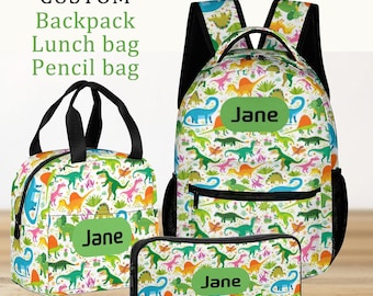 May include: A custom backpack, lunch bag, and pencil bag set. Each item features a white background with a colourful dinosaur pattern. The backpack and lunch bag have black accents. The name "Jane" is printed on each item.