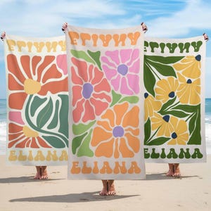 Op de afbeelding: Drie strandlakens met bloemenpatronen in oranje, roze en geel. Elk laken heeft de naam "ELIANA" onderaan en het woord "FRIYAY" bovenaan in een retro lettertype. De lakens worden vastgehouden op een zandstrand met de oceaan op de achtergrond.