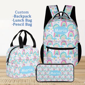 May include: A custom backpack, lunch bag, and pencil bag set. Each item features a white background with a repeating pattern of colourful rainbows, clouds, and hearts. The name "Maria" is printed on each item in a blue oval.