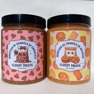 Op de afbeelding: Twee potten Cloudy Treats granola, één met chocolade en de andere met koekjes. De potten hebben zwarte deksels en labels met schattige illustraties en de tekst "200 gram". De chocoladepot heeft een roze achtergrond en de koekjespot heeft een gele achtergrond.