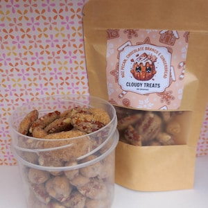 Könnte beinhalten: Ein durchsichtiger Plastikbehälter und ein brauner Papiersack gefüllt mit gezuckerten Pekannüssen. Der Beutel trägt ein Etikett mit der Aufschrift "Cloudy Treats" und "Noz Pecan, Chocolate Branco e Gingerbread". Der Hintergrund ist ein Blumenmuster.