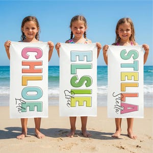 May include: Three white beach towels with colorful names: "CHLOE", "ELSIE", and "STELLA". Each name is in a different color and font, with a smaller name below. The towels are held by three young girls on a sandy beach with the ocean in the background.